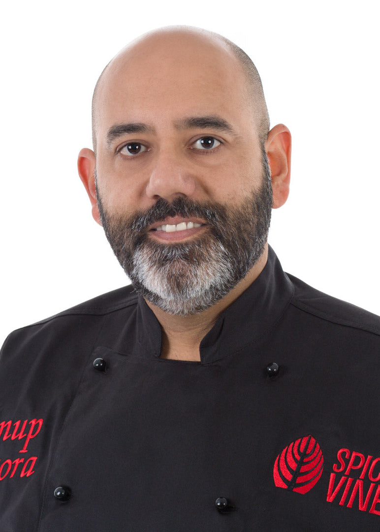 A handsome man with a bald head and salt and pepper  beard wearing a black chef jacket with red embroidered text reading 'Anup Arora' and 'SPICE VINE', photographed against a plain white background.